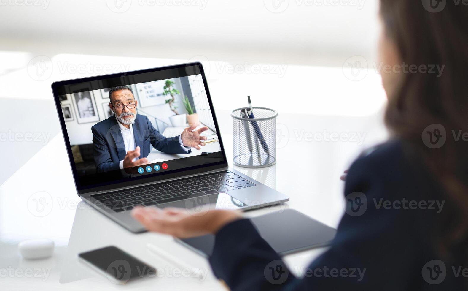 A businesswoman sits at her desk, engaged in a call with a colleague on her laptop. She has a serious expression on her face, listening intently to the man on the screen photo