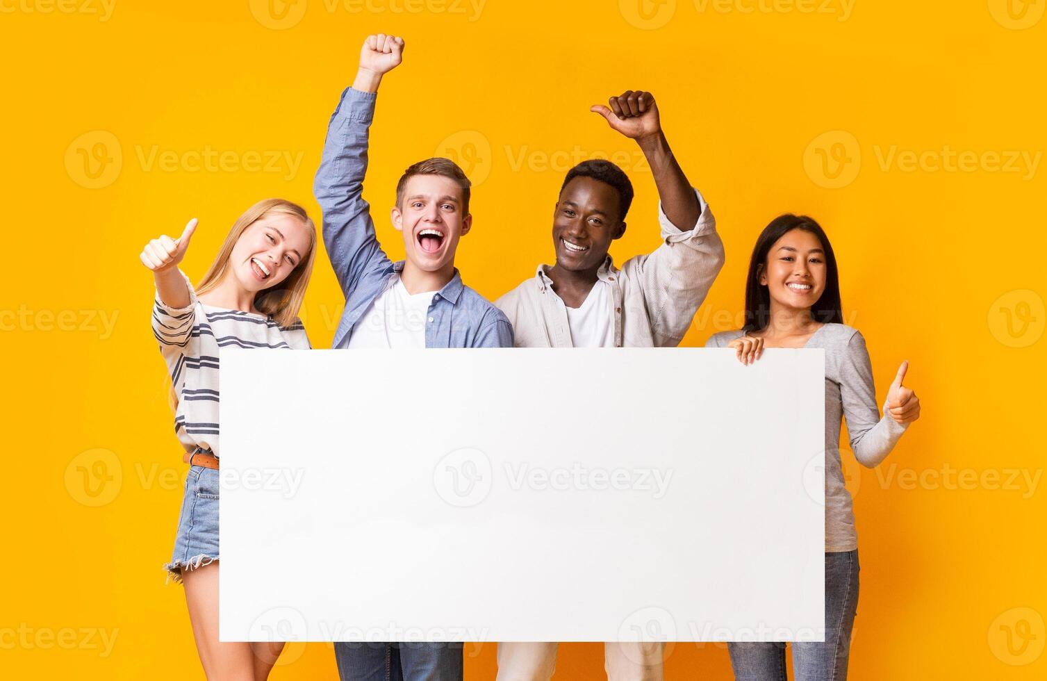 Joyful multiracial teenagers with empty billboard showing thumbs up over yellow background photo