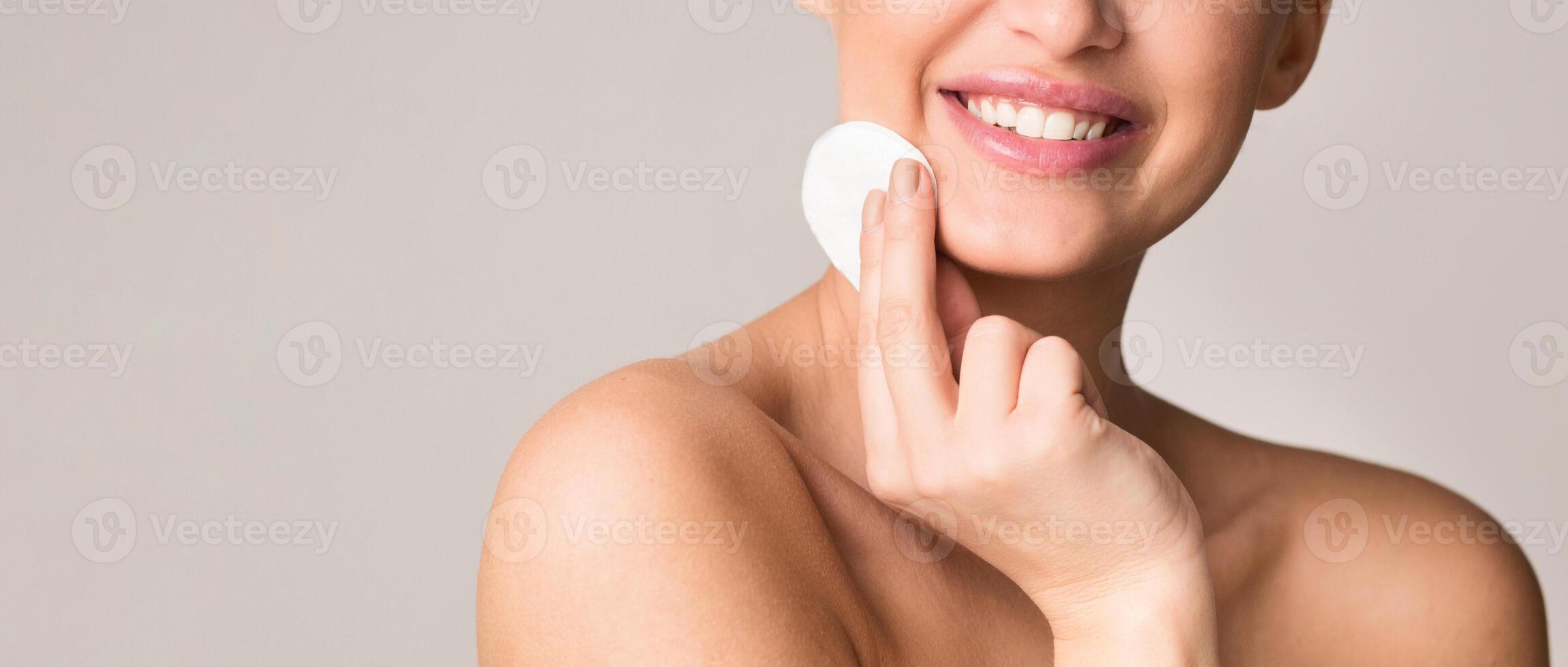 A smiling woman uses a cotton pad to clean her face in a well-lit room. She appears content and happy, focusing on her skincare routine, cropped. copy space photo