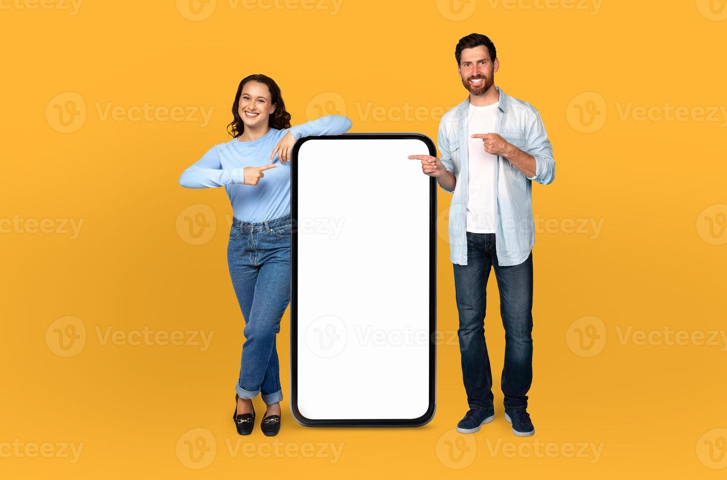 A man and woman are standing in front of a blank screen, both pointing towards it with their index fingers. The background is a solid yellow color, creating a vivid contrast with the neutral screen. photo