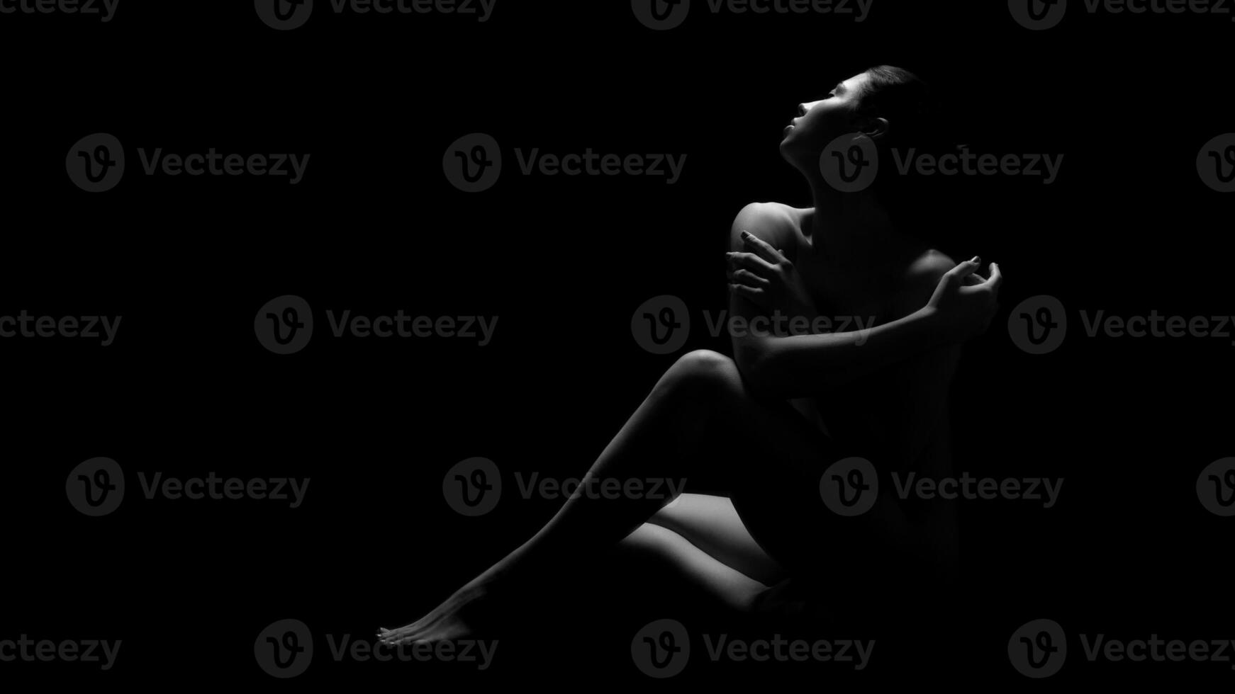 A black and white photograph of a woman sitting in darkness, illuminated only by a single light source. She has her arms crossed, and her gaze is directed upwards. photo