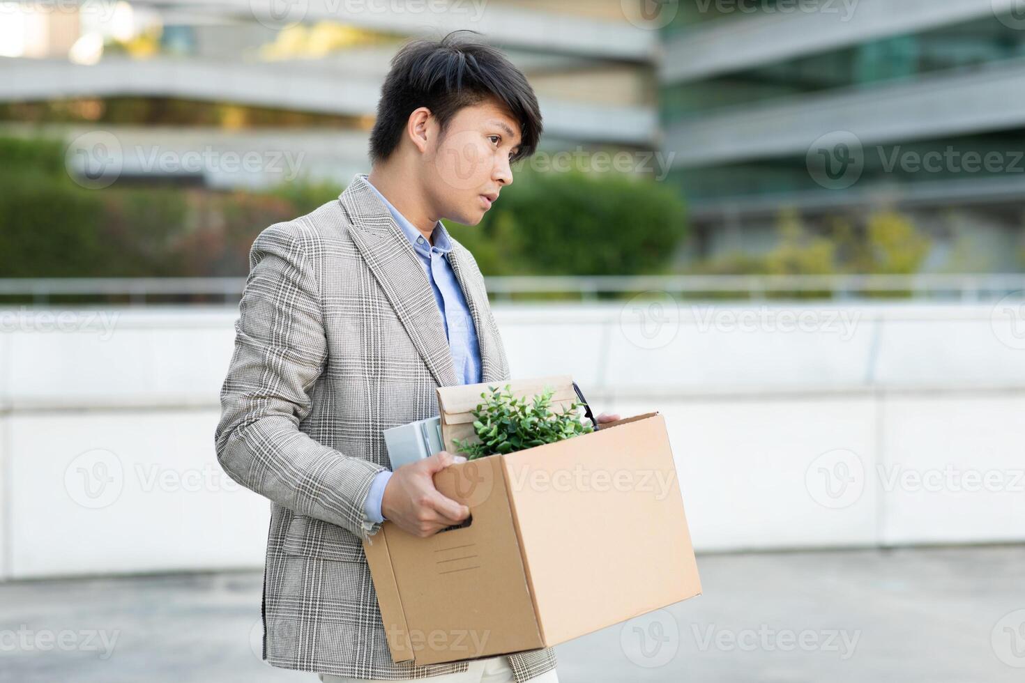 A young Asian man walks away from a modern office building, carrying a cardboard box filled with personal items. He appears dejected and downtrodden, perhaps having just been laid off or resigned photo