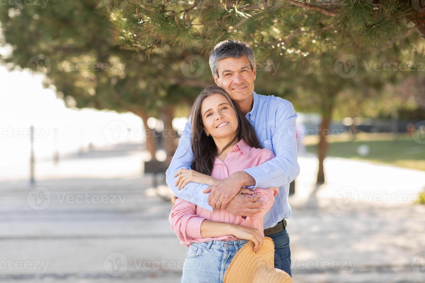 A happy couple stands close together on a beach promenade, surrounded by trees, embracing each other with smiles. The sun shines brightly, creating a warm atmosphere during their outing. photo