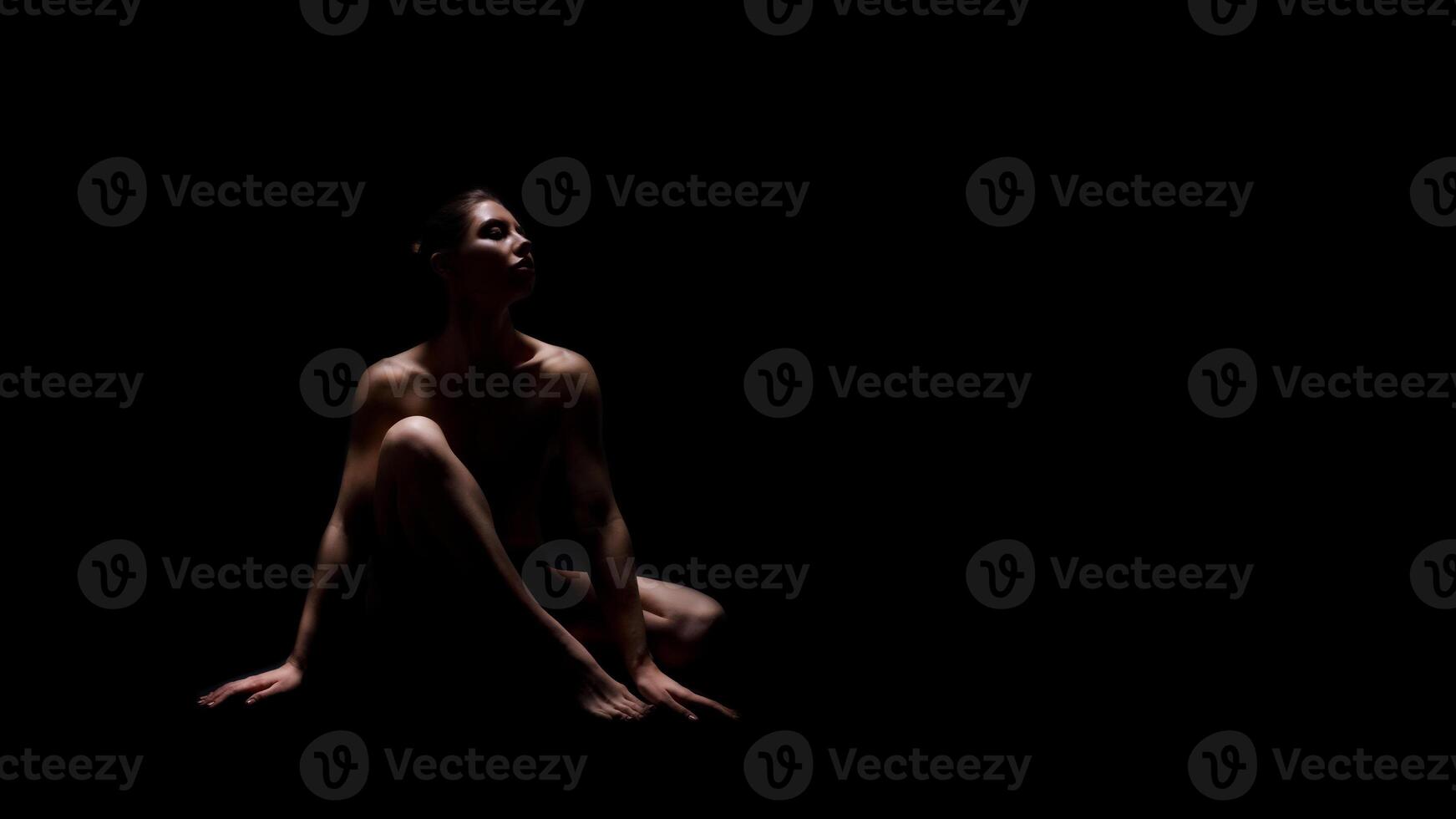A woman sits in a dark room with only a single light source highlighting her body. The light creates a dramatic effect, illuminating her form and leaving her face and features in shadow, copy space photo