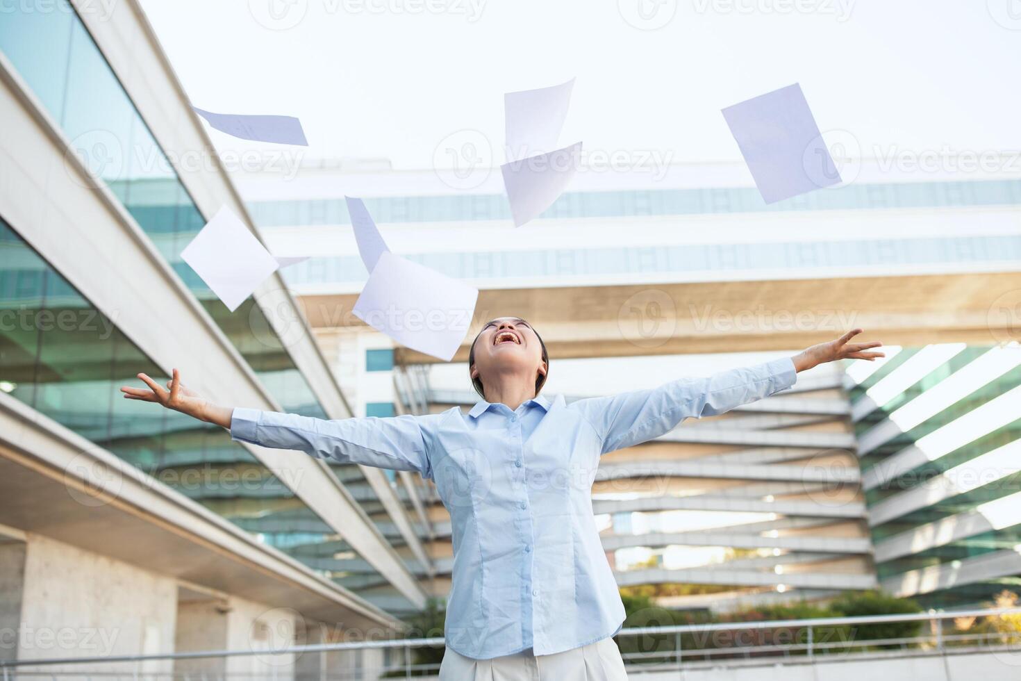 Chinese woman is energetically tossing papers into the air outside an office building. The scene is filled with flying papers against the backdrop of a corporate skyscraper. photo