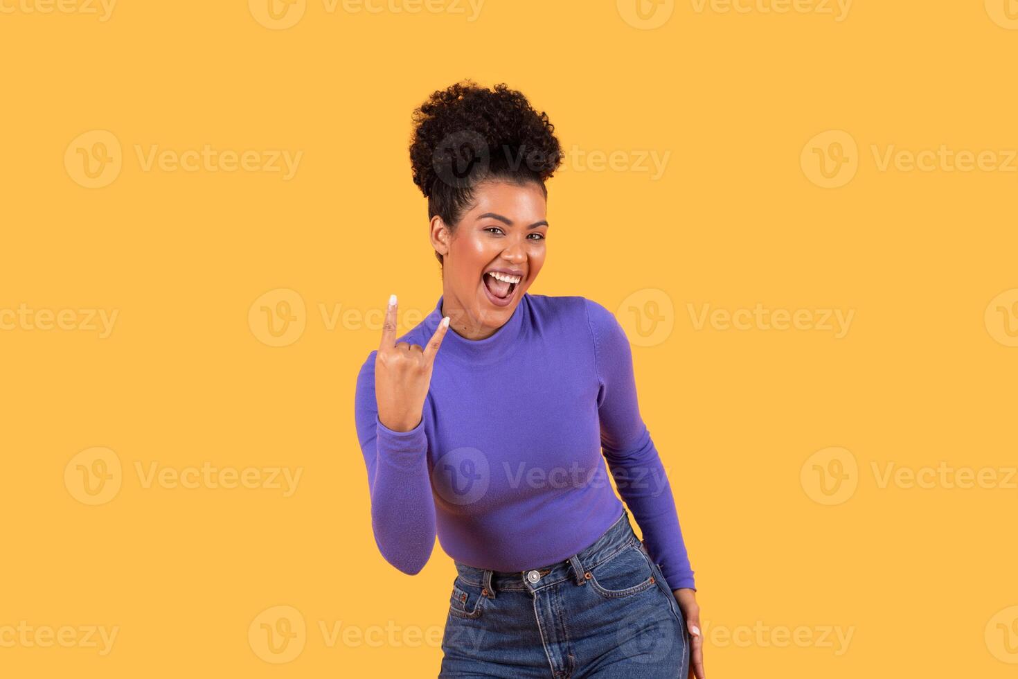A young woman stands confidently, flashing a rock hand gesture with a big smile on her face. She is wearing a fitted purple sweater paired with high-waisted jeans. photo