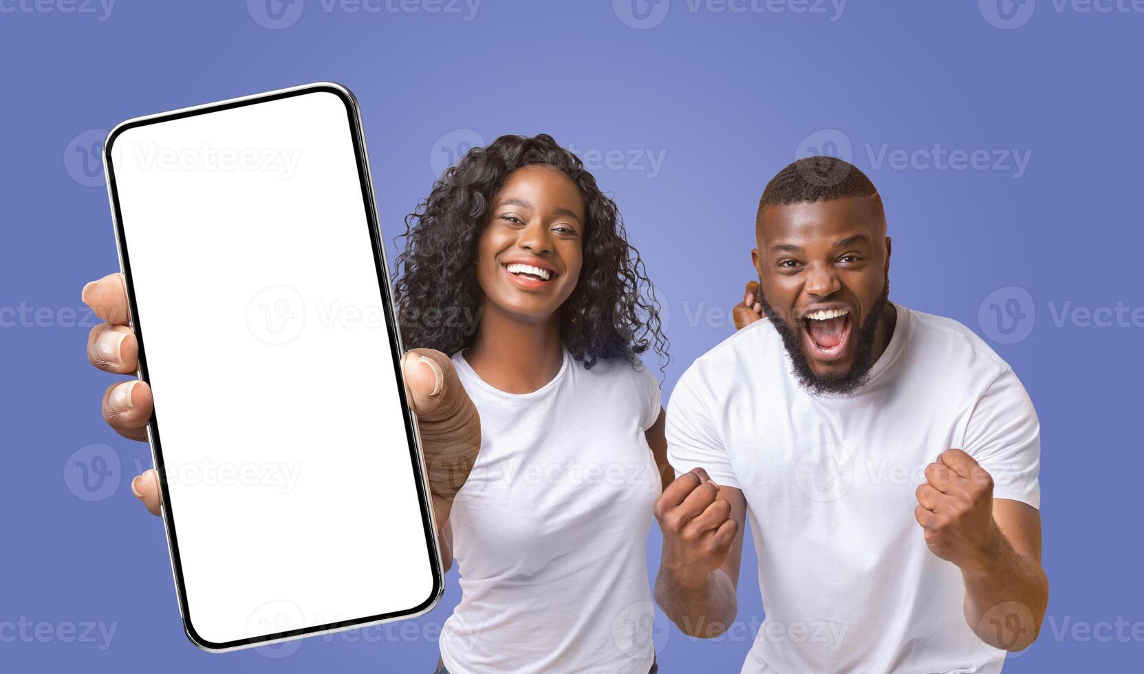 A young black couple is excited about something they are looking at on a smartphone. The woman is pointing at the blank screen while the man has his fists pumped in the air, indicating excitement photo
