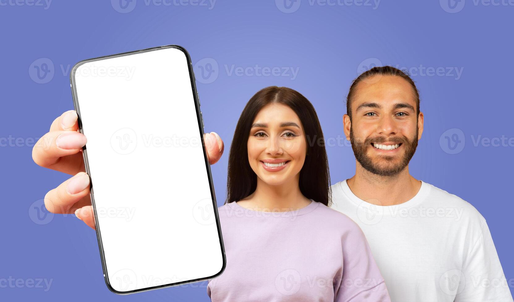 A young woman holds a smartphone with a blank screen in front of her and a young man, both smiling, standing behind her. The background is a solid blue color. photo