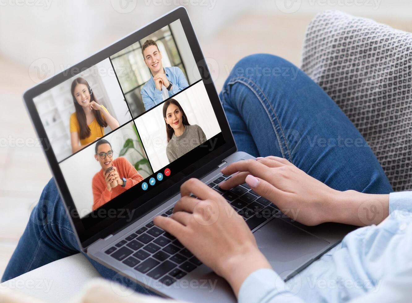A woman sits on a couch and uses a laptop computer for a conference. The laptop screen displays four participants in separate frames, cropped photo
