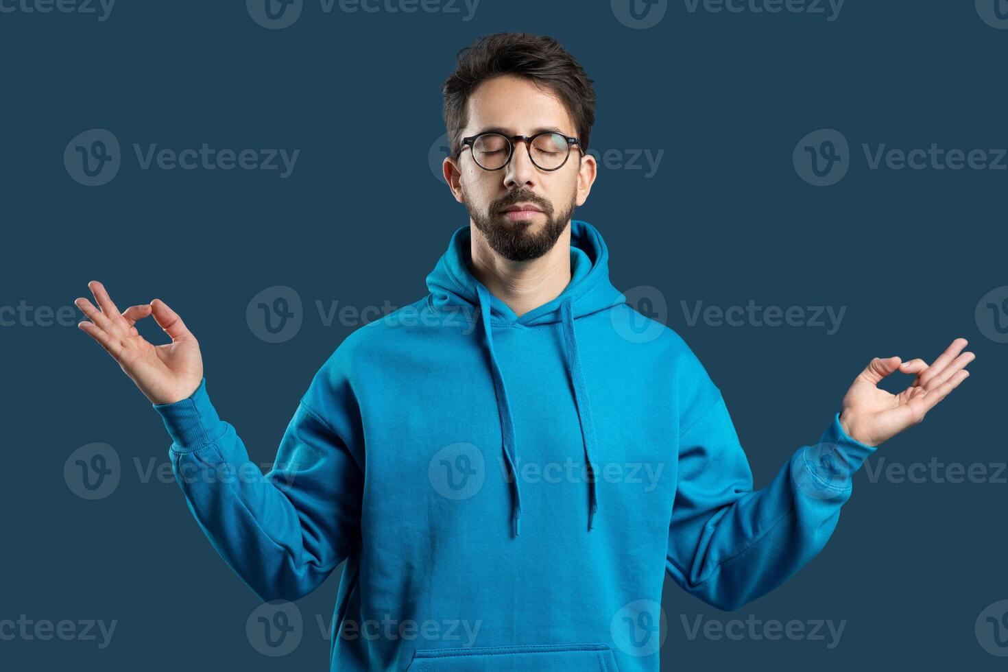 A young man with a beard and glasses, wearing a bright blue hoodie, stands against blue background with his eyes closed in a peaceful posture, practicing a meditative pose with his hands gently raised photo