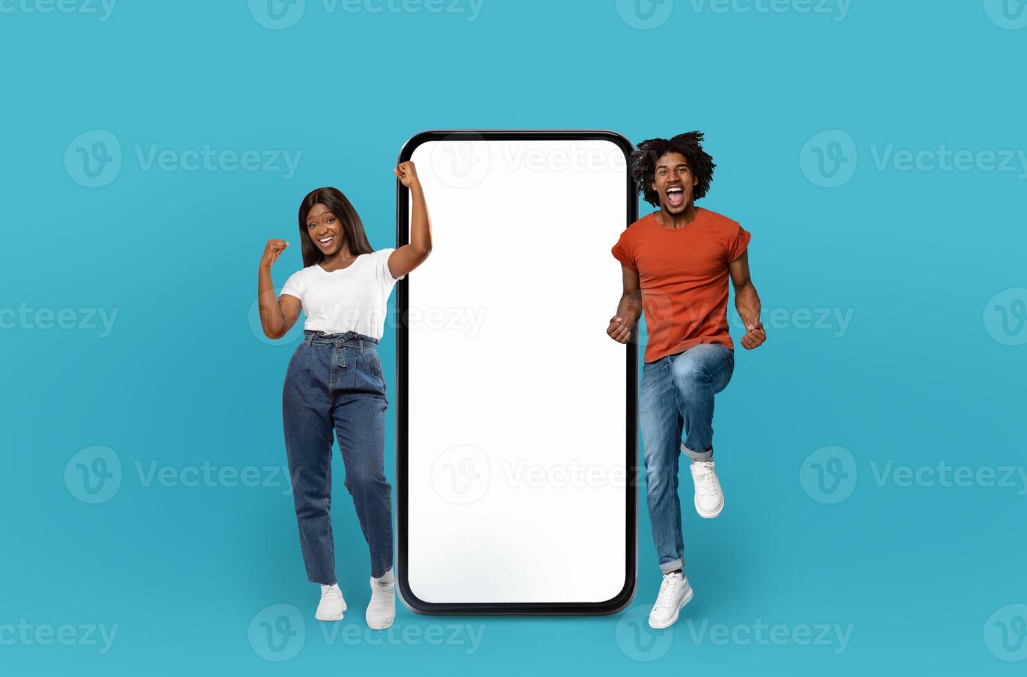 Two African American individuals are energetically jumping up and down while holding a blank screen against a solid blue background. photo