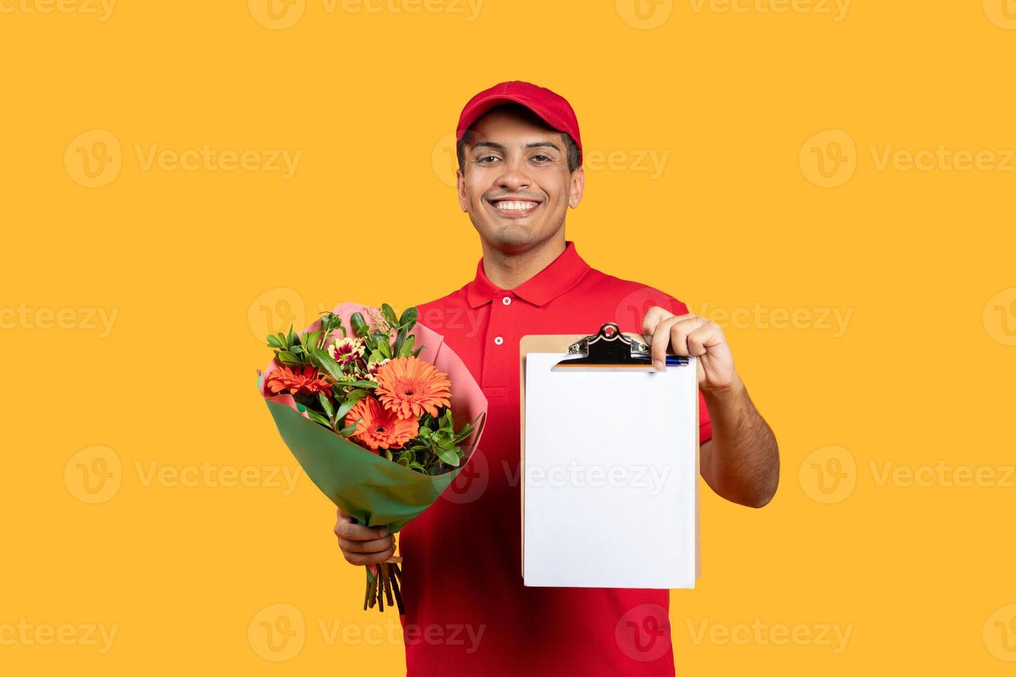 A smiling delivery man, wearing a red uniform and cap, holds a beautiful flower bouquet in one hand and a clipboard in the other, standing against a vibrant yellow background. photo