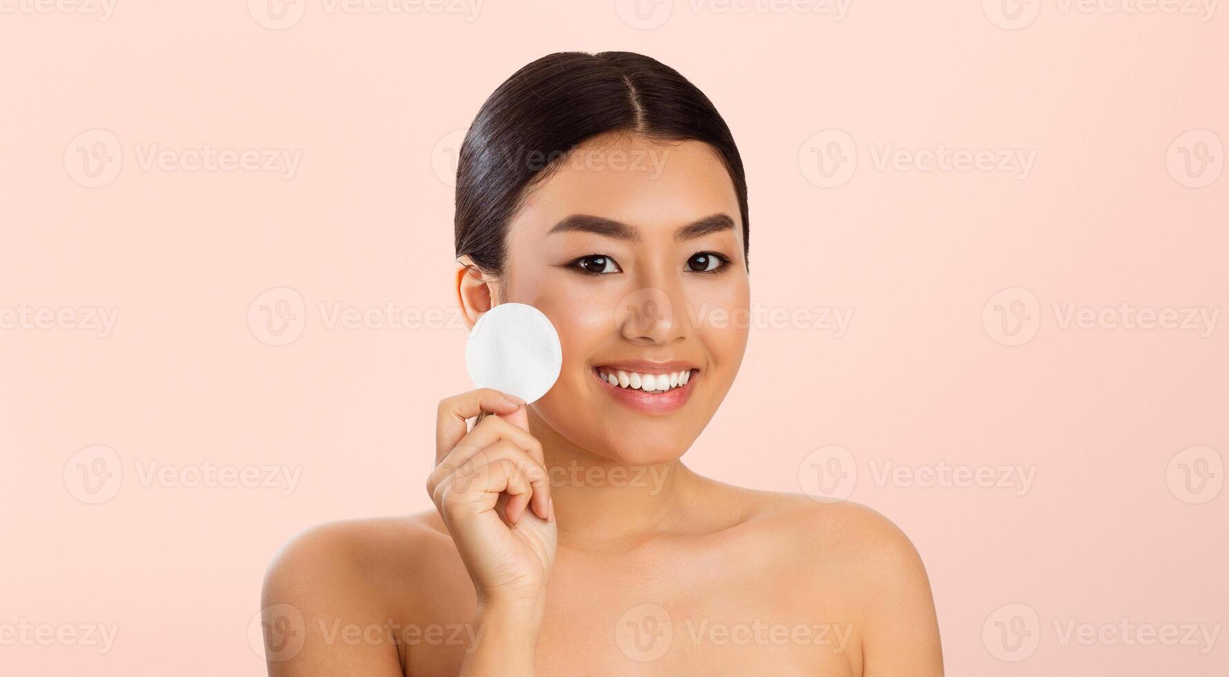 Asian woman with dark hair and a bright smile holds a white cotton round against her cheek. She is looking directly at the camera and has a light pink background behind her. photo