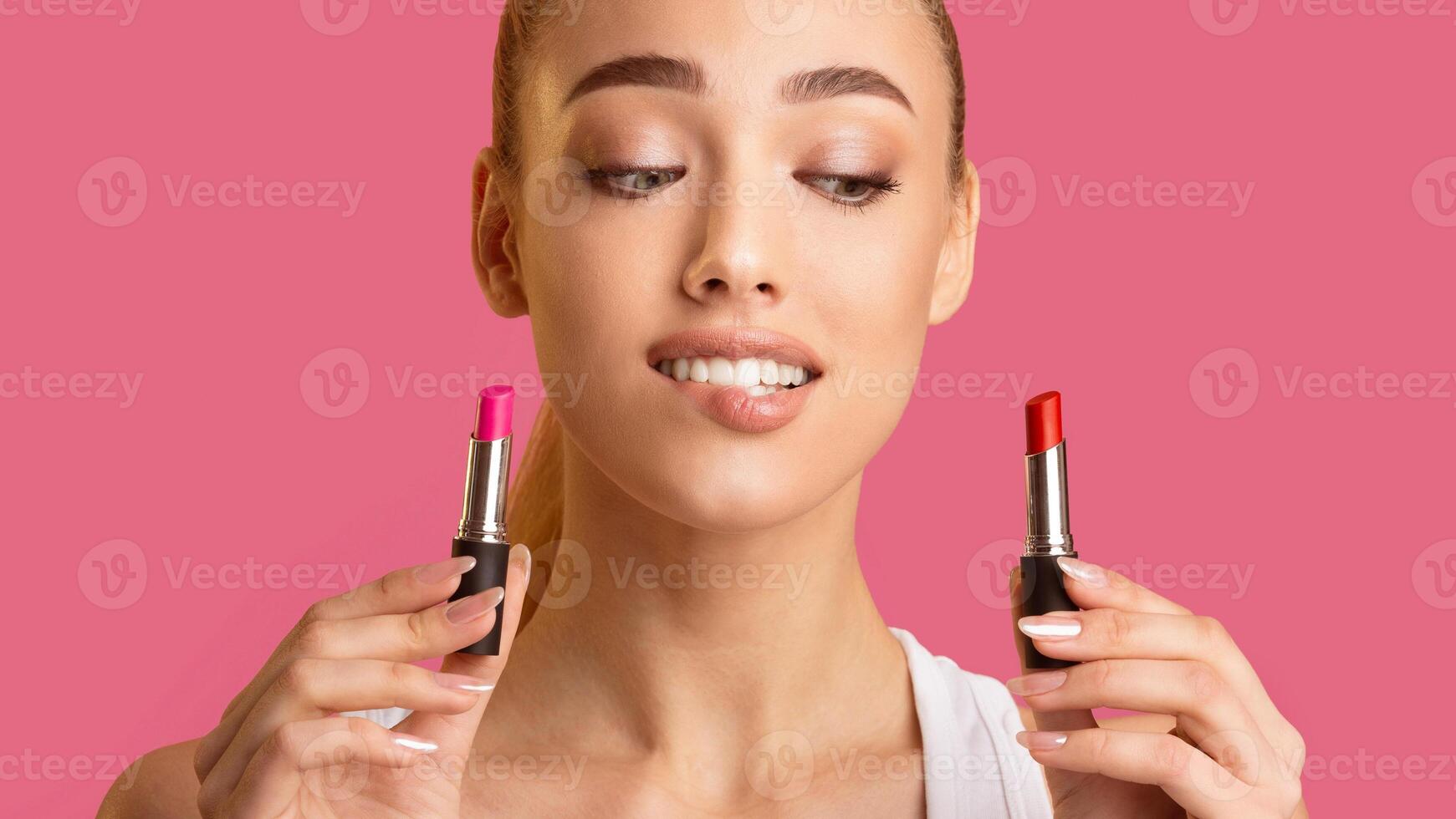 A woman holds a pink lipstick in her left hand and a red lipstick in her right hand. She is looking at the lipsticks thoughtfully. The woman is standing in front of a pink background. photo