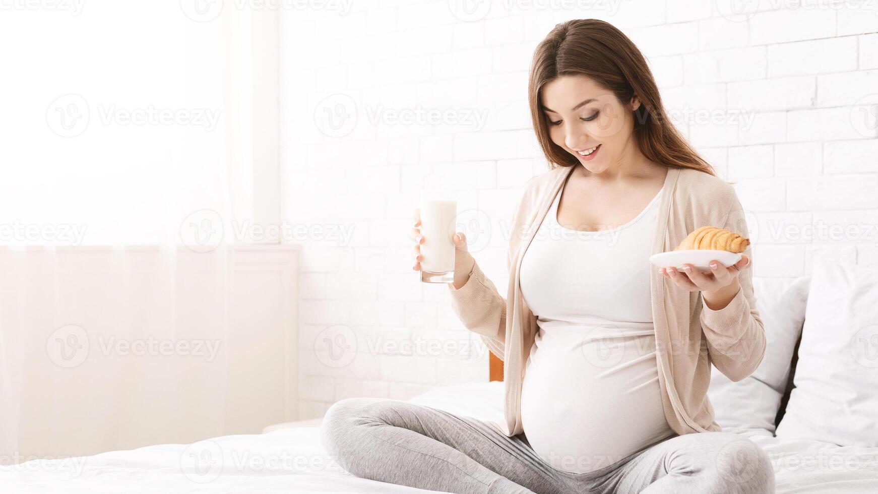 Young pregnant woman having brunch of milk and croissant, sitting on bed and looking at belly, free space photo