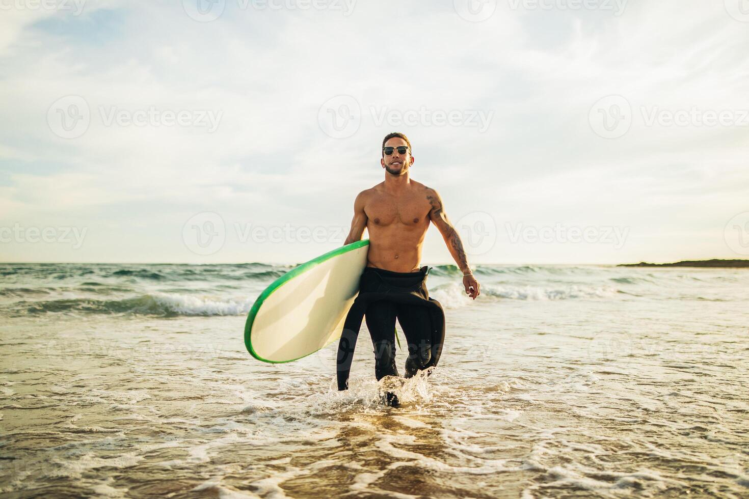 A man wearing a wetsuit and sunglasses walks out of the ocean with a surfboard under his arm. The sun is setting in the background, casting a warm glow on the water. photo
