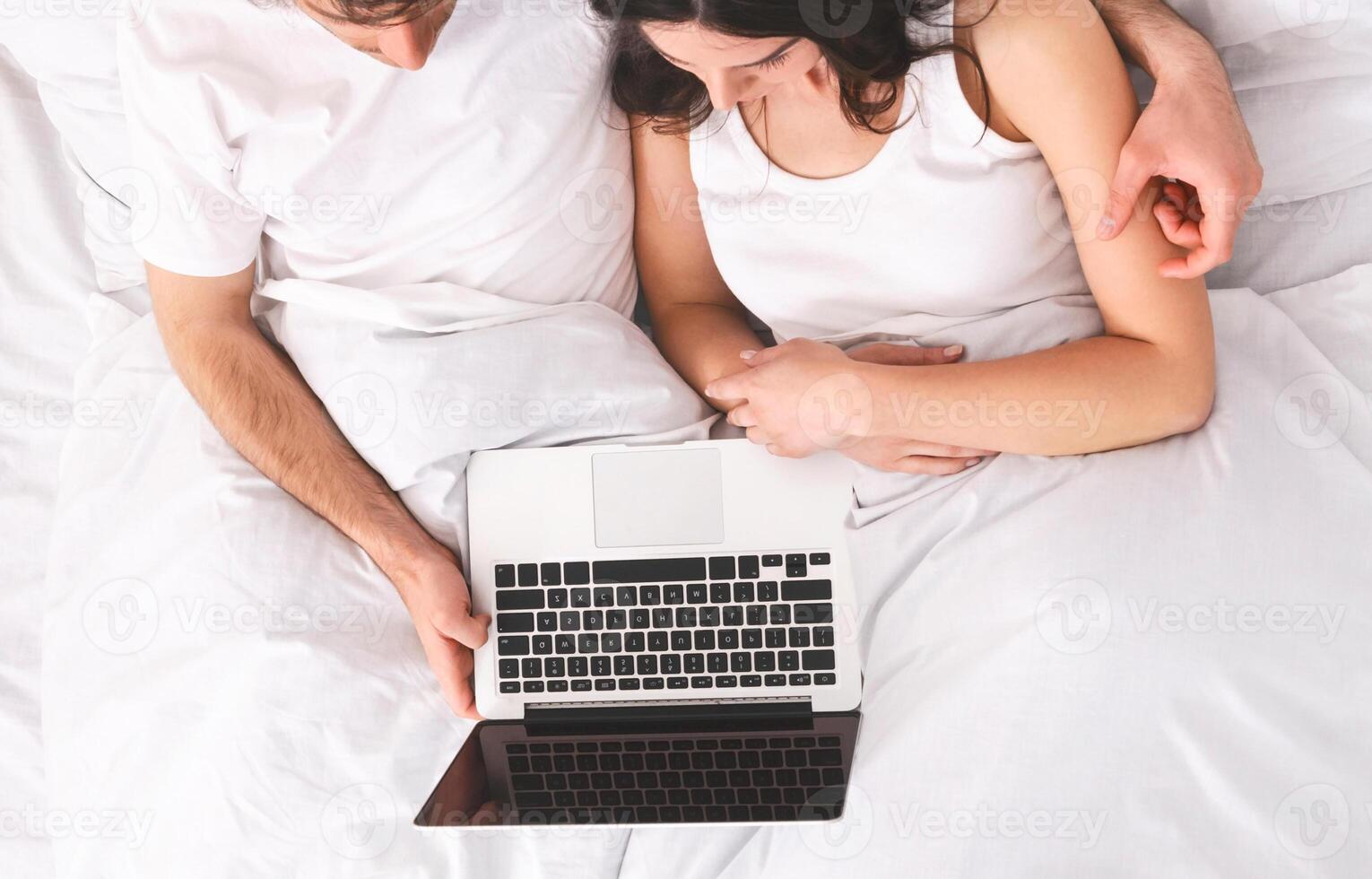 A couple in bed, the woman is lying down, the man is sitting up and holding a laptop. They are both looking at the laptop screen. The image is taken from an overhead angle. photo