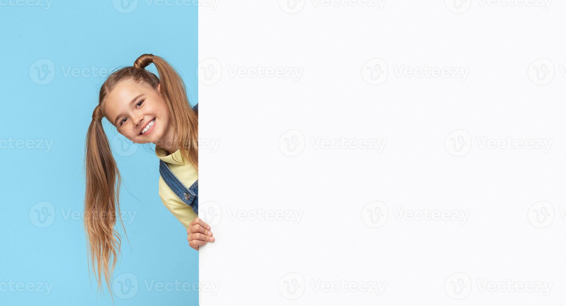 A young girl with long blonde hair in pigtails peeks around a white board, smiling at the camera. She is wearing a yellow shirt and blue overalls. The background is a solid light blue. photo