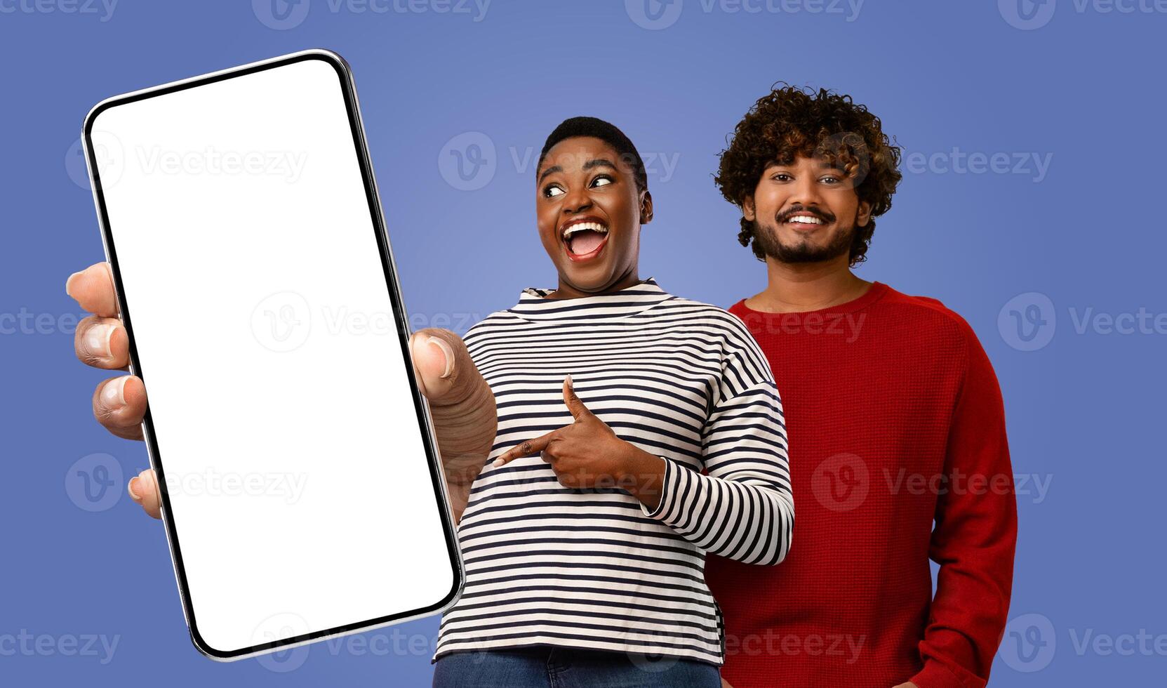 Black woman and Indian man stand in front of a blue background. They are both looking at the phone that the woman is holding. photo