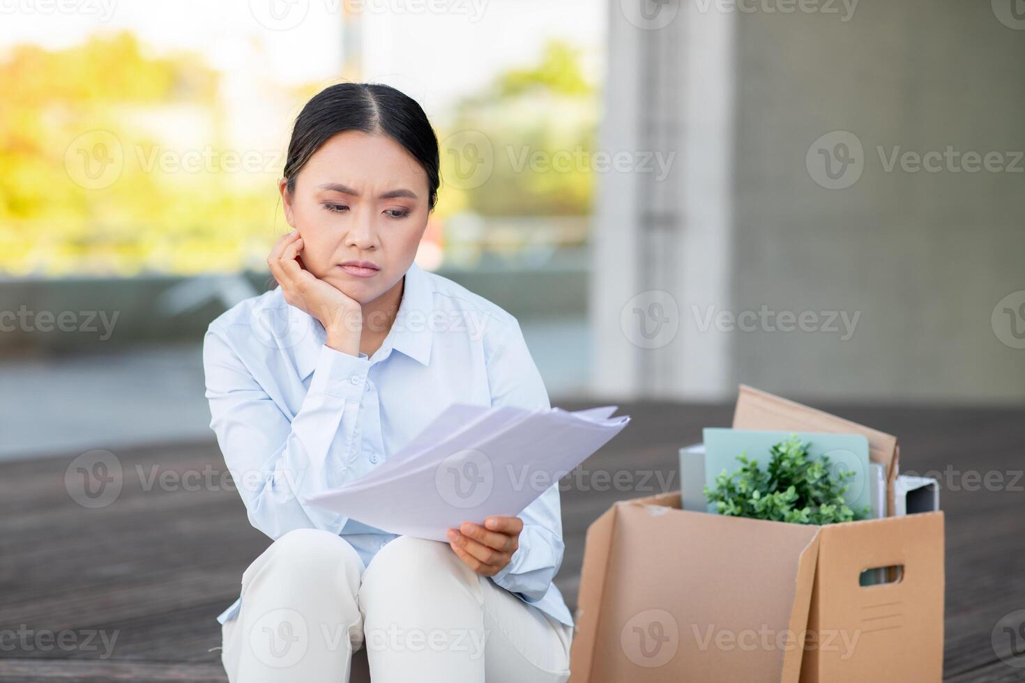 un joven profesional asiático dama aparece profundo en pensamiento como ella comentarios documentos relacionado a trabajo aplicaciones ella se sienta en pasos en un moderno urbano configuración, con un cartulina caja conteniendo suministros foto