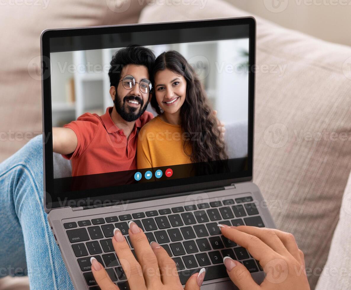 A woman sits on a couch and chats with Indian couple on her laptop. The woman is smiling and typing on the keyboard. The couple on the screen is also smiling. photo