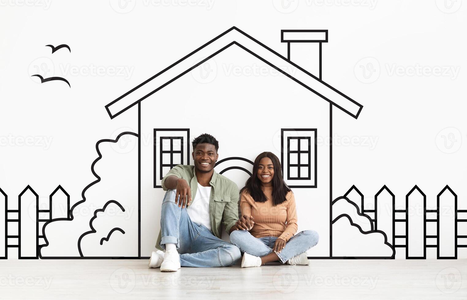 African American Spouses are seated on the floor, facing each other, with a drawing of a house propped up in front of them. They appear to be discussing or admiring the sketch. photo