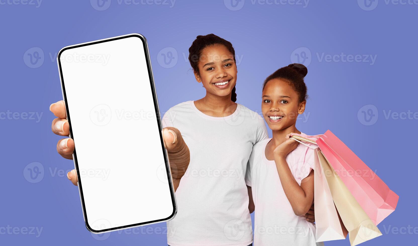 Two happy black girls, one holding a smartphone with a blank screen and the other holding a stack of shopping bags, stand in front of a solid purple background photo