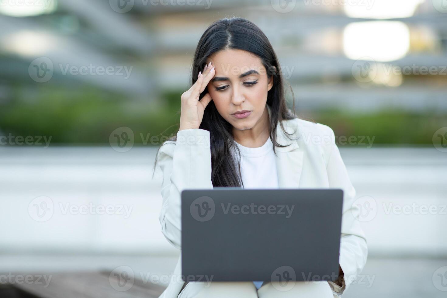 A young woman in a white blazer sits outdoors, using a laptop computer. Her expression is serious and focused, and she appears to be concentrating on the screen. photo