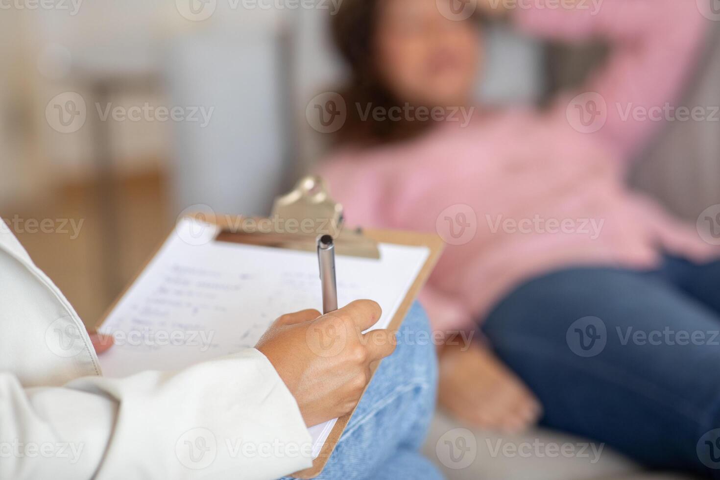 The image shows a therapist taking notes during a session with a client. Therapists hand is holding a pen and writing on a clipboard. Client is out of focus in the background, reclining on a couch photo