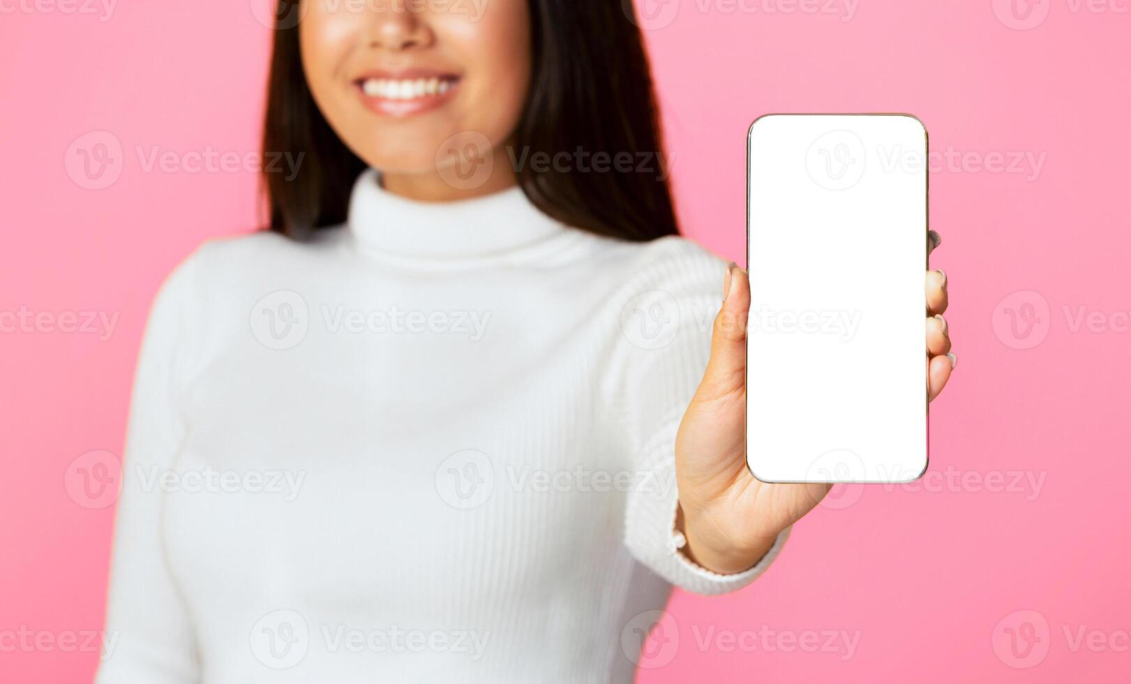 A young woman in a white turtleneck sweater smiles and holds out a smartphone with a blank white screen in front of a pink background, cropped photo