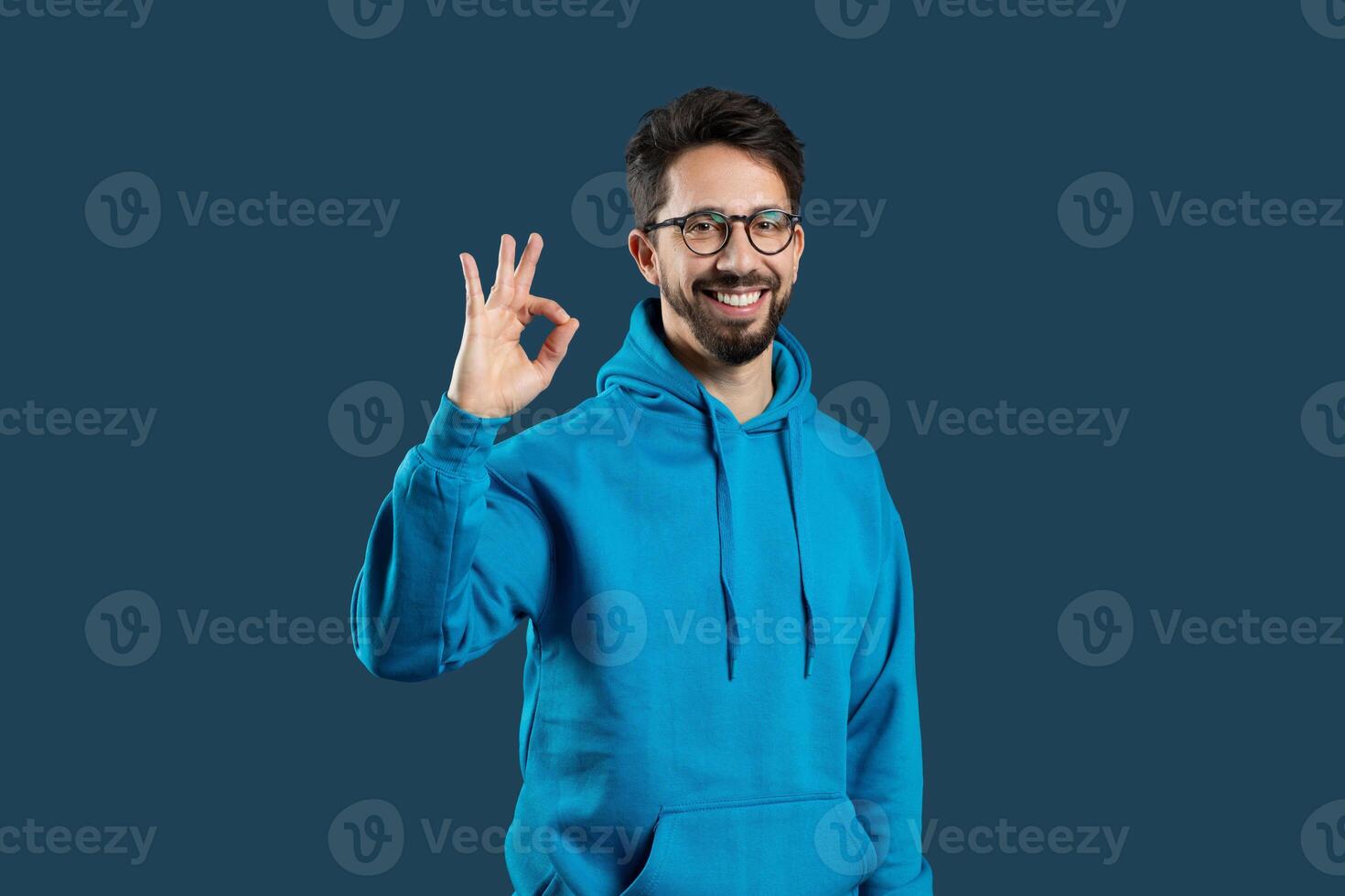 A cheerful young man with glasses and a beard is wearing a bright blue hoodie and making the OK hand gesture, standing in front of a dark blue backdrop with a broad, friendly smile. photo