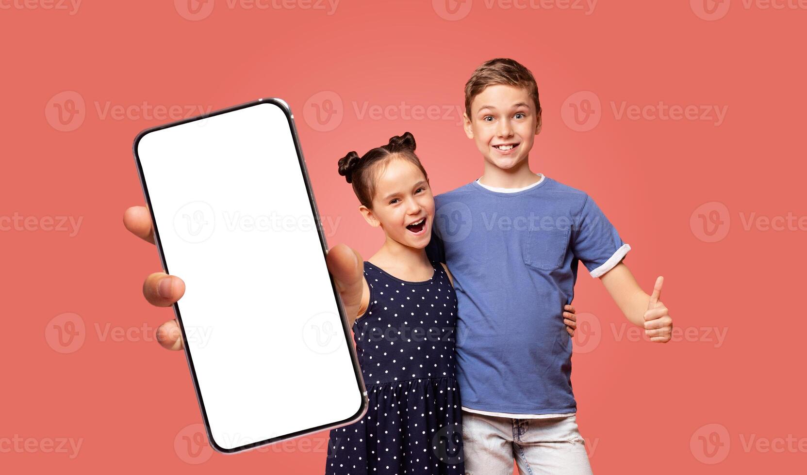 Two children, a boy and a girl, stand in front of a pink background. The boy is giving a thumbs up. The girl is holding a smartphone with a blank white screen. photo