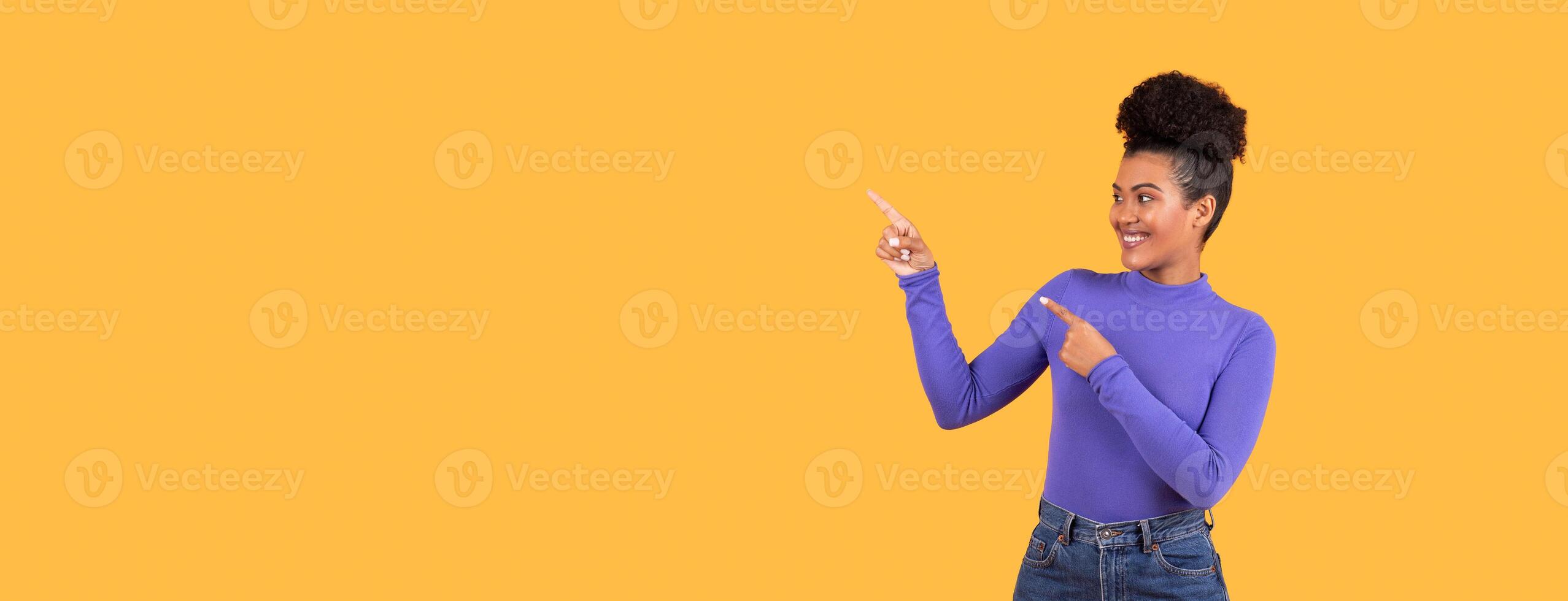 A young woman with curly hair is enthusiastically pointing to copy space, wearing a purple long-sleeved top and high-waisted jeans. The vibrant yellow background adds to the cheerful atmosphere photo