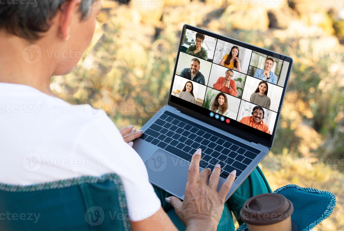 A senior woman is participating in a conference call on a laptop computer while sitting outdoors on a green chair. The laptop screen displays multiple feeds of other people photo