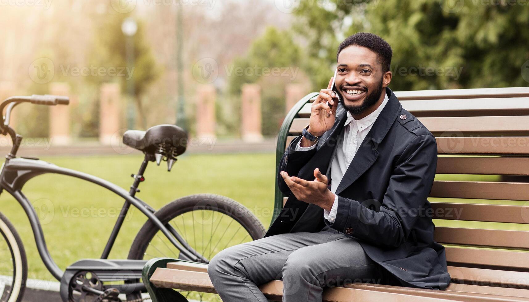 Black business professional is using his phone in a park, seated on a bench with a bicycle beside him. The scene is calm and serene, highlighting a balance between work and nature. photo