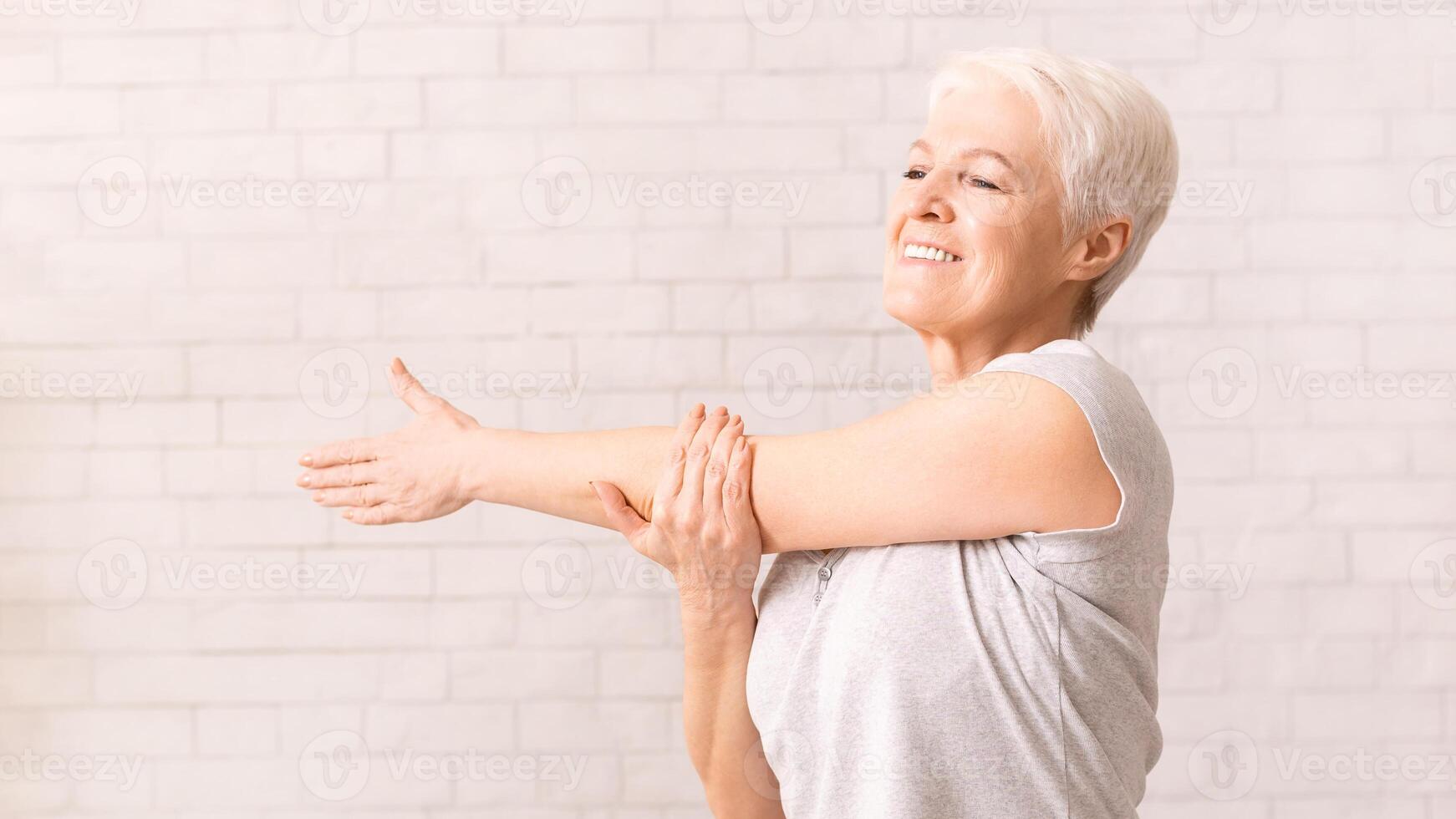 A joyful elderly woman with short white hair is engaging in a light stretching routine, extending her arms across her chest and smiling widely photo