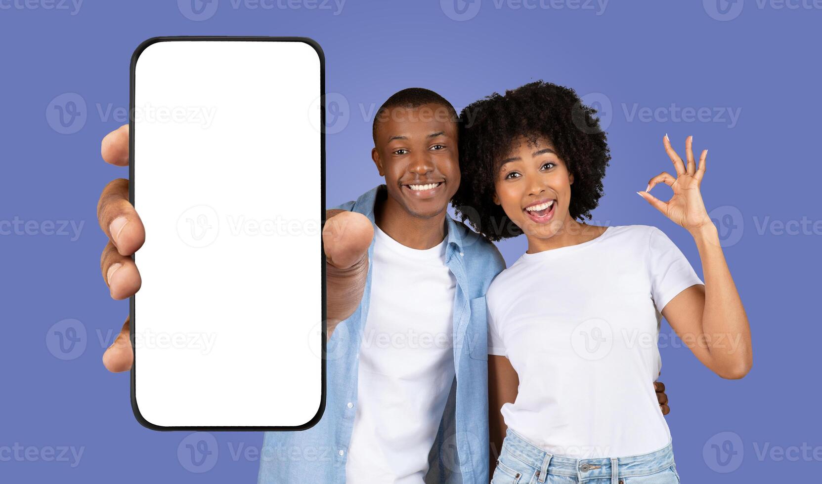 A young African American couple stands together in front of a purple background, smiling and looking at the camera. The man is holding a smartphone with a blank screen, woman showing okay photo