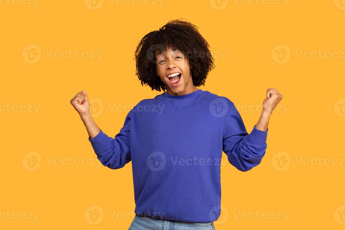 This photo shows a happy young man with an curly hairstyle, wearing a blue sweatshirt, celebrating his success with a big smile and raised fists. He is standing in front of yellow background