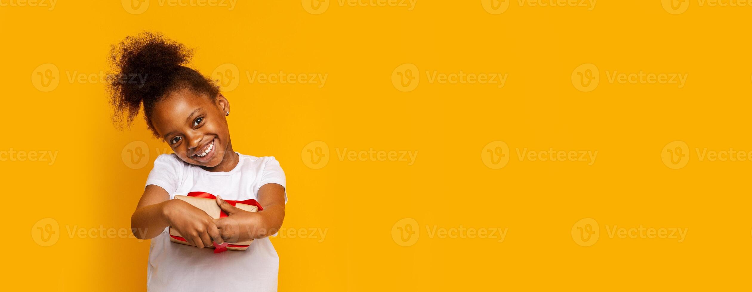 A young black girl stands cheerfully in front of a vivid yellow backdrop, holding gift. Her joyful expression radiates happiness as she engages with the camera, creating a playful atmosphere. photo