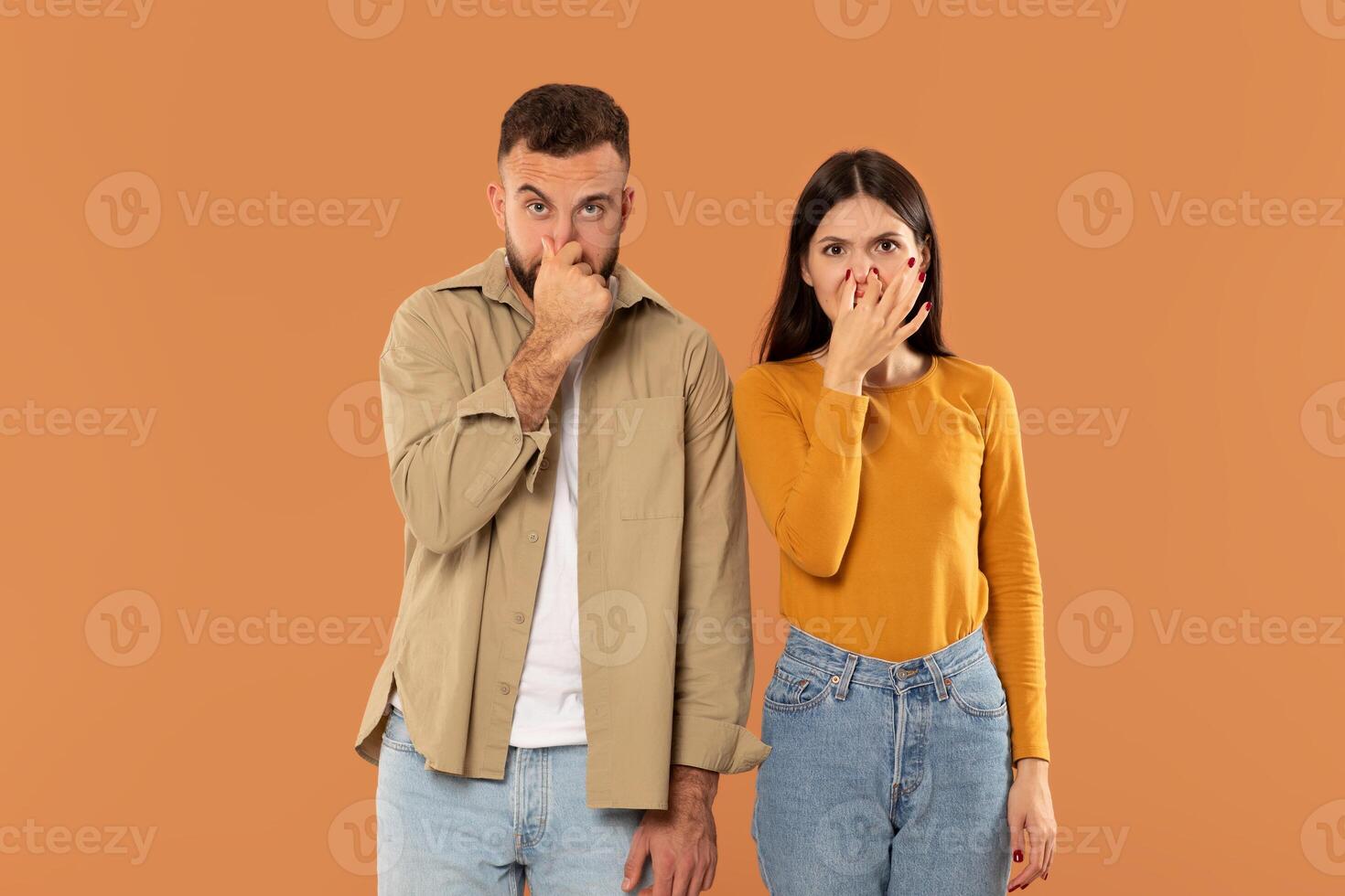 A man and a woman stand against an orange background with their hands covering their noses. Both are casually dressed photo