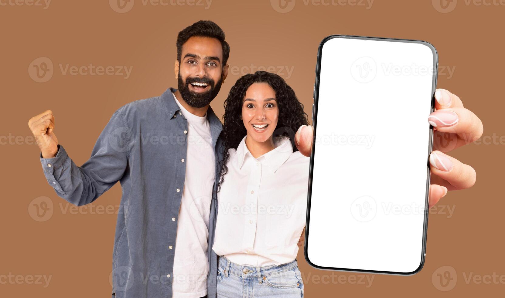 A happy multiracial couple is standing in front of a brown background, holding a large smartphone with a blank screen. photo