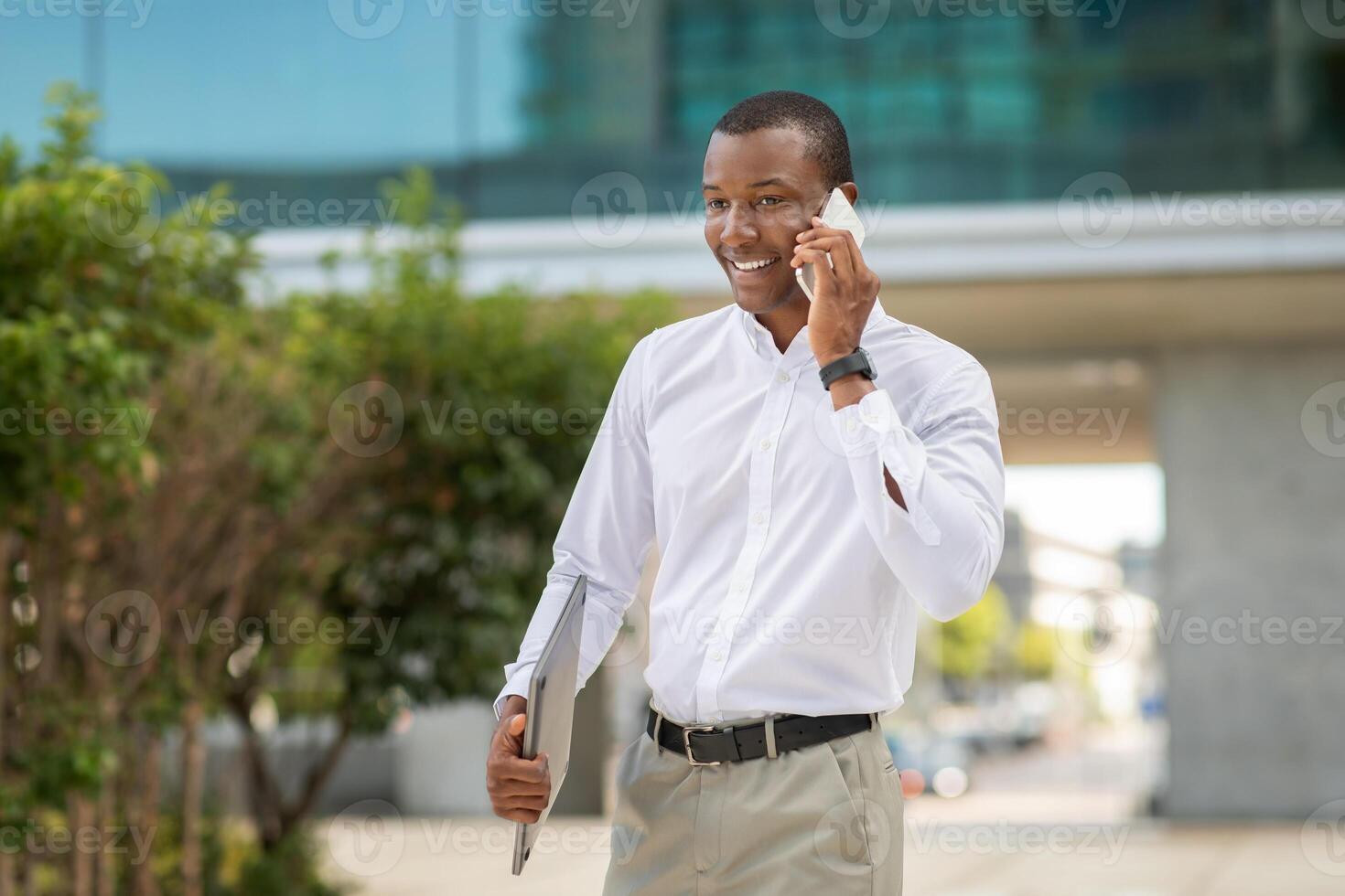 African American man in business attire is talking on his phone while standing outside an office building. He appears confident and engaged, holding a laptop under his arm, copy space photo