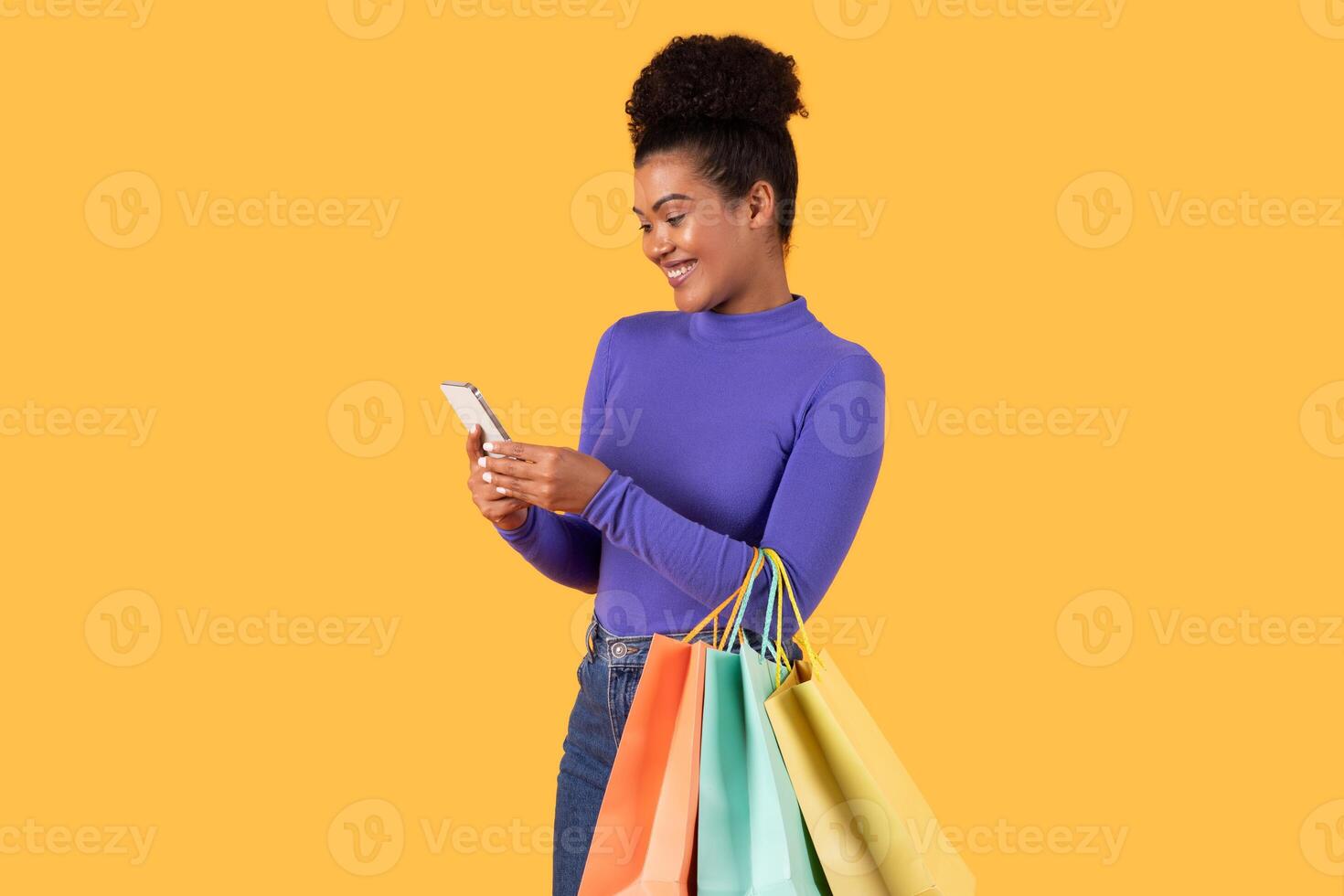 A cheerful woman stands holding several vibrant shopping bags in one hand while using her smartphone with the other. She appears to be engaged with her device, showcasing a modern shopping photo