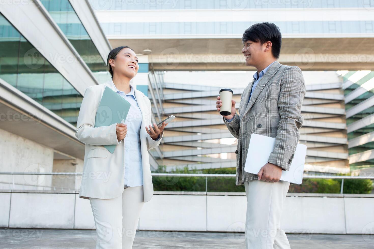 Two Asian business professionals, dressed in formal attire, are having a friendly conversation while standing outside a contemporary office building. photo