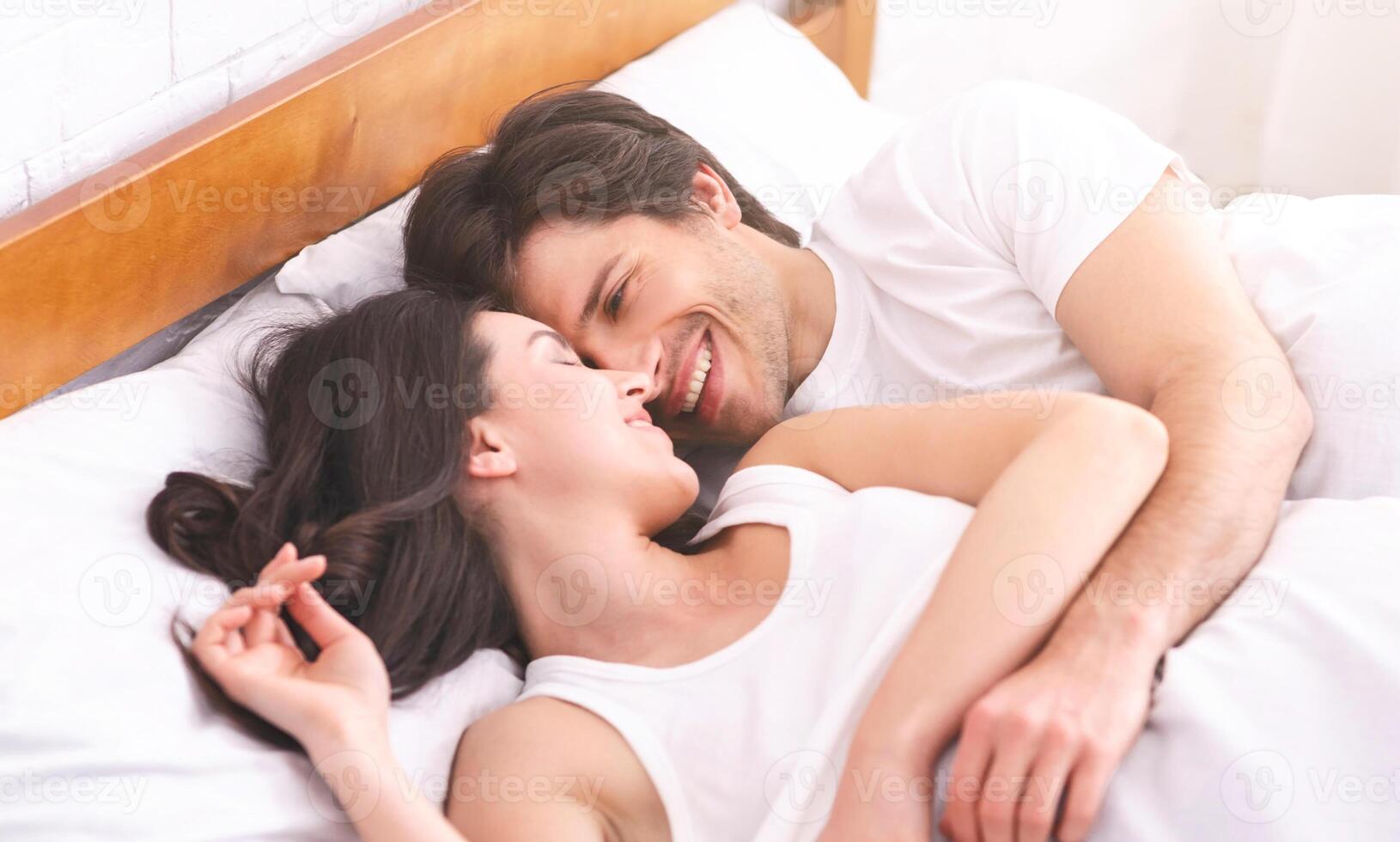 A couple lies in bed, a man and a woman, both smiling and looking at each other. The woman lies on her back with her head resting on the pillow, while the man is on his side, looking down at her photo