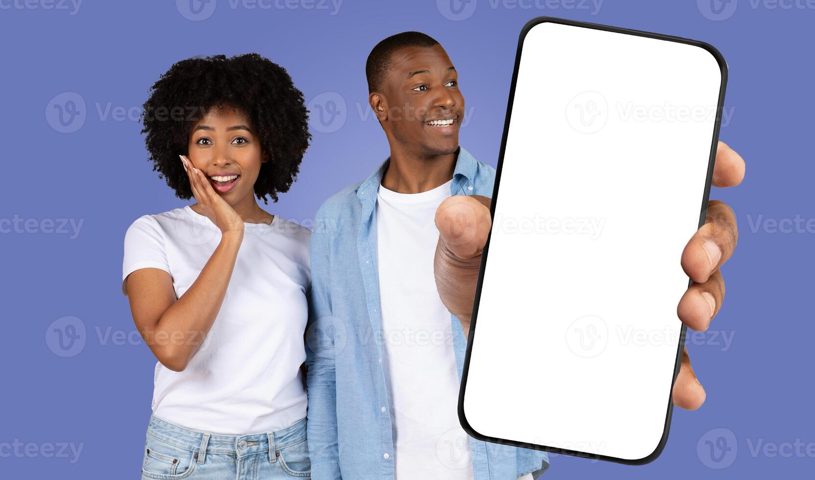 African American young woman looks surprised while a man holds a phone with a blank screen. The background is a solid blue color. photo