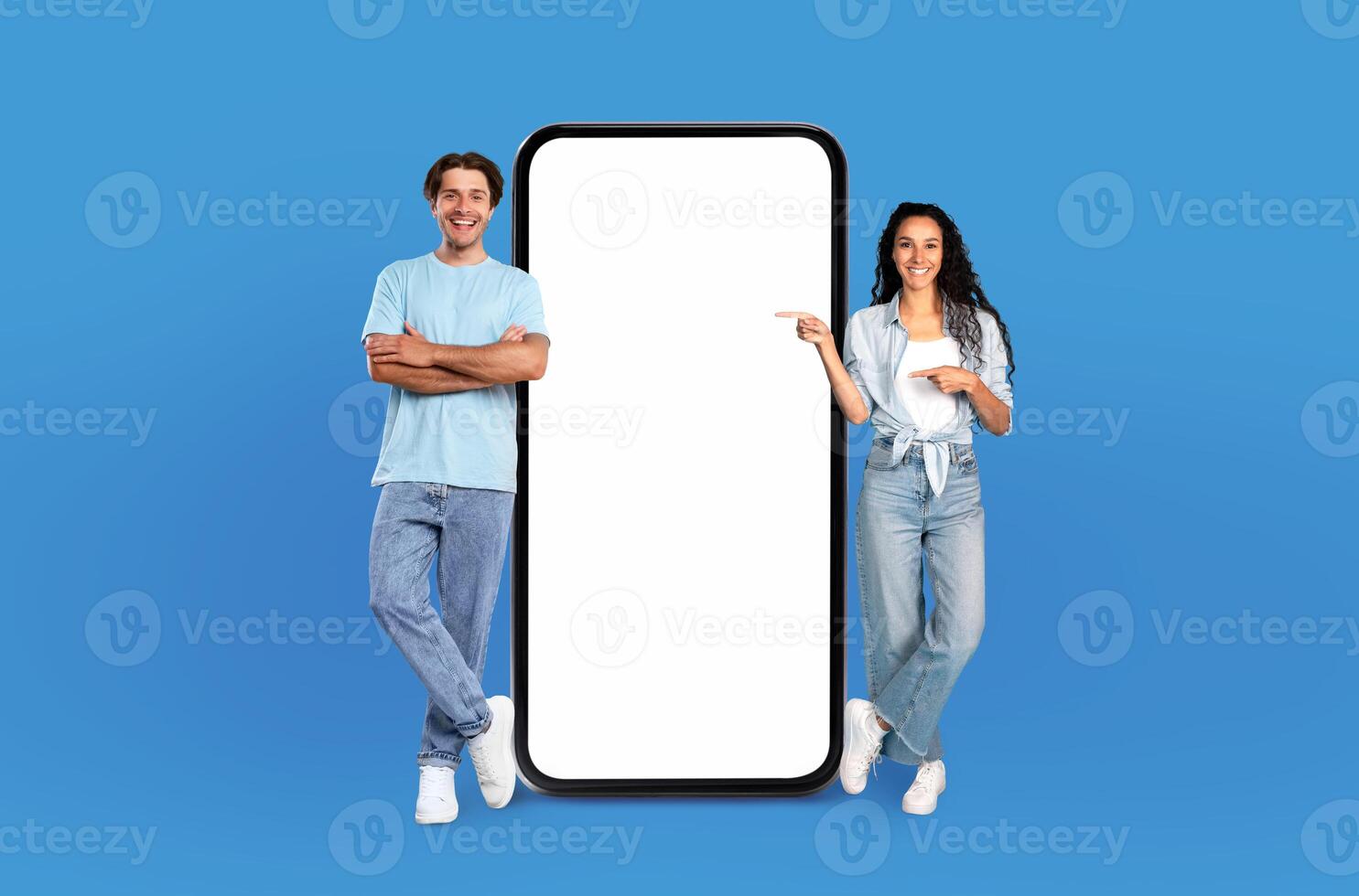 A cheerful young man and woman stand side by side against a vibrant blue backdrop, confidently presenting a gigantic, blank vertical smartphone screen between them, mockup photo