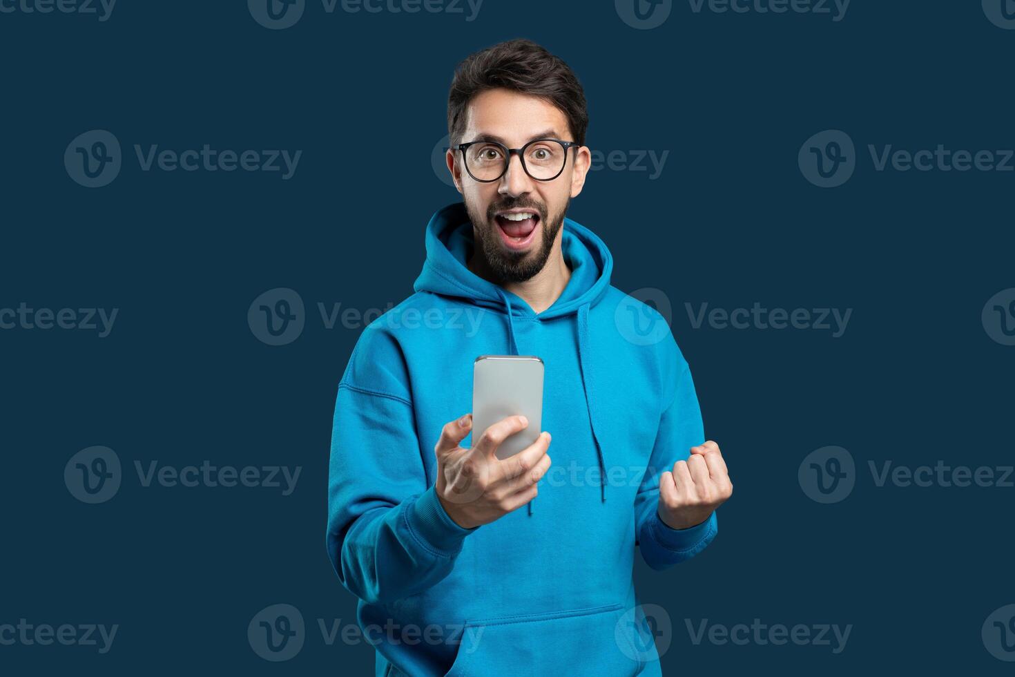 A man wearing a blue hoodie is standing outdoors, holding a cell phone in his hand. He appears to be looking at the screen intently, possibly texting or browsing, gesturing photo