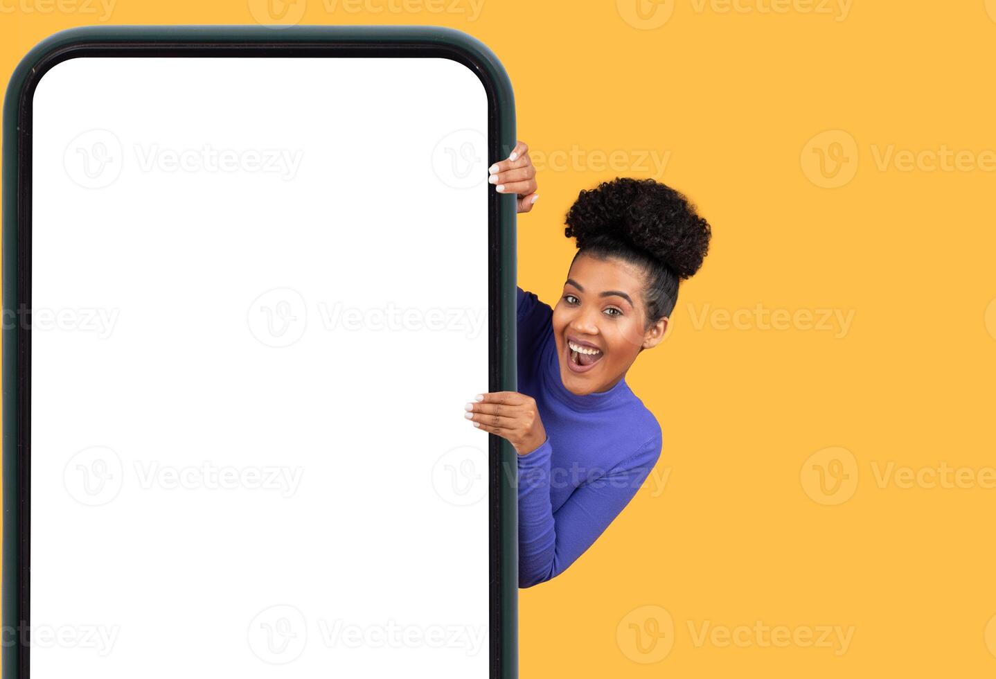 A young woman with curly hair joyfully leans out from behind a large mobile device, showcasing her excitement. The bright yellow backdrop adds a cheerful touch, enhancing the playful atmosphere photo