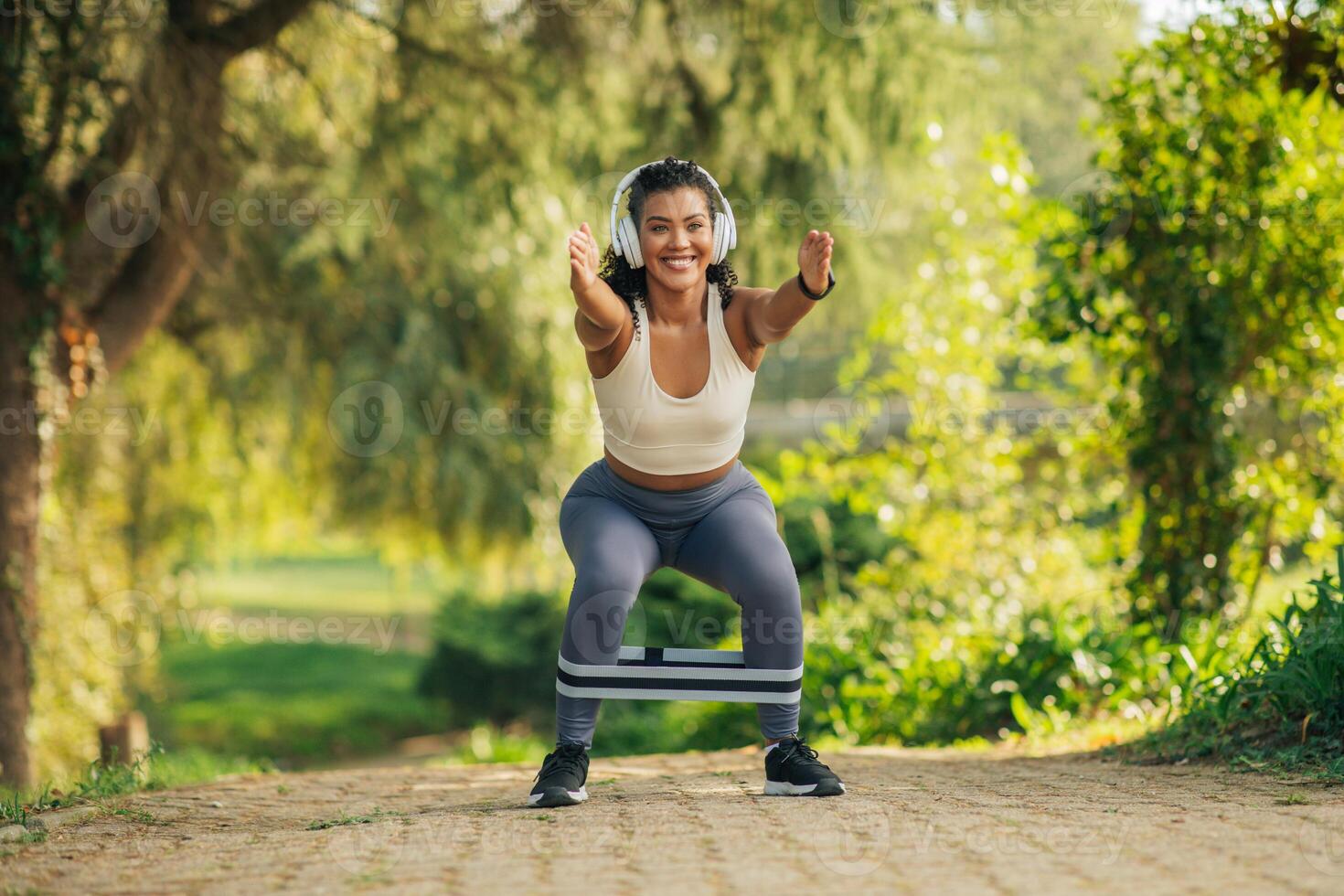 A woman wearing headphones, a white sports bra, gray leggings, and black running shoes, performs a squat exercise with a resistance band in a park. photo